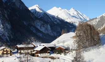 A picturesque mountain landscape in winter with snow-covered peaks and a small village in the valley. The clear, blue sky adds a serene atmosphere.