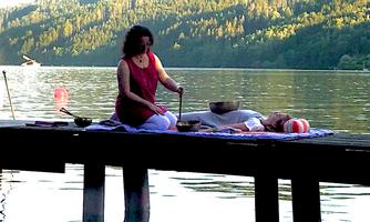 A woman is sitting on a dock by the water and playing a sound bowl. In the background, gentle hills and trees are visible.