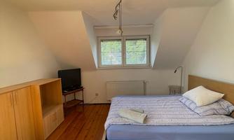 A cozy bedroom with wooden flooring and a large window. There is a bed, a television, and a desk.