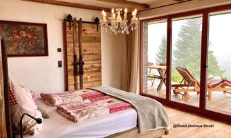 A cozy bedroom with a large bed and rustic wooden furniture.  
Large windows offer a view of nature and a comfortable seating area on the terrace.