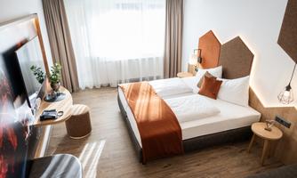 A modern hotel room with a double bed, a cozy seating area, and a large window. The decor is in warm colors and creates an inviting atmosphere.
