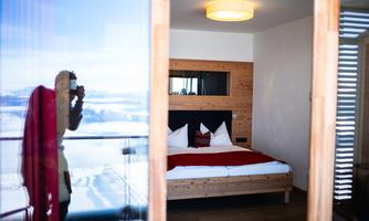 A modern hotel room with a double bed and light wood. In the background, a reflective glass surface can be seen.