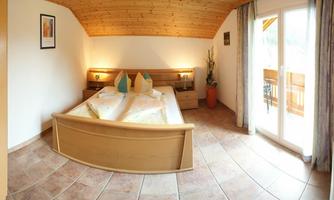 A cozy bedroom with a large bed and wooden ceiling. Large windows provide access to a balcony and plenty of daylight.