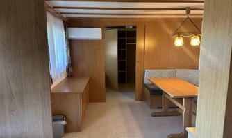 A simple living room with wooden walls and a table. There is a dining area with a bench and a ceiling lamp.