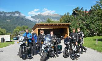 A group of motorcyclists stands in front of their motorcycles in a picturesque landscape. In the background, mountains and a blue sky can be seen.