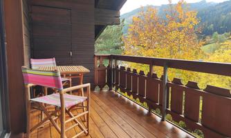 A cozy balcony with two chairs and a wooden table. In the background, autumn trees and mountains can be seen.