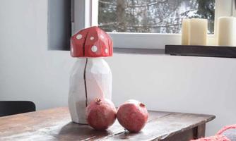 A table with a rustic wooden surface, on which a decorative vase in the shape of a mushroom stands. Two apples are lying beside it, and in the background, you can see a snow-covered landscape through the window.