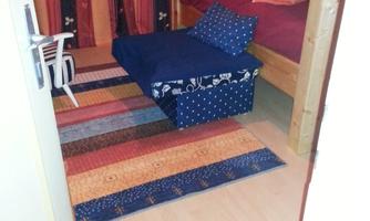 A cozy room with colorful rugs and a bed. On the left side, there is a small armchair and extra blankets are placed on the bed.
