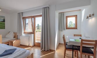 A bright room with large windows and wooden flooring. There is a table with chairs and a cozy bed.
