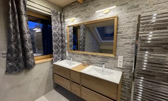 A modern bathroom with two sinks and a large mirror. The walls are decorated with stone tiles and large windows provide daylight.