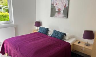 A cozy bedroom with a large bed and a purple blanket. A painting of flowers hangs on the wall, and there are two lamps on the nightstands.