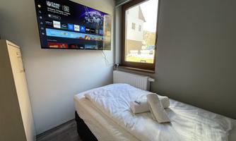 A simple bedroom with a single bed and a flat-screen TV on the wall. The window provides natural light and a view outside.