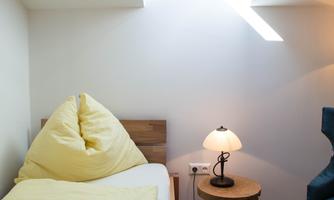 A cozy bedroom with a single bed and yellow bedding. Next to the bed is a small nightstand with a lamp and a blue armchair in the corner.