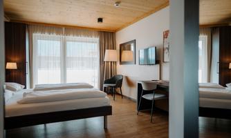 A modern room with two beds, a television, and a table. Large windows provide plenty of daylight.