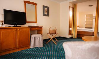 A cozy hotel room with a TV, a desk, and a mirror. In the background, another room with a bed can be seen.