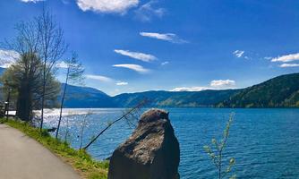 A calm lake, surrounded by gentle hills and trees. The sky is blue with some clouds.