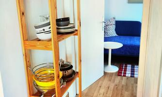 A narrow wooden shelf stands in the hallway and is loaded with dishes. In the background, there is a cozy living area with a blue couch and a small table.