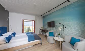 A modern hotel room with a large bed and blue-white accents.  
There are comfortable armchairs and a TV mounted on the wall, as well as a nice view through the window.