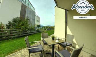 A cozy terrace with a table and chairs, surrounded by greenery. In the background, a modern building can be seen.