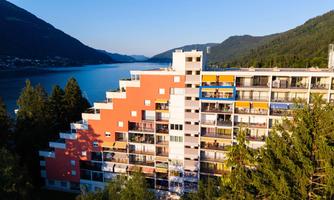 A modern building with colorful balconies stands by the shore of a lake. In the background, mountains and a clear sky can be seen.