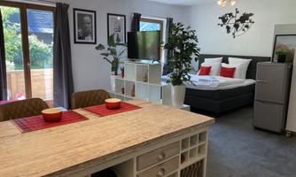 A cozy living room with a modern kitchen and a dining area. In the background, there is a bedroom with a double bed and a TV.