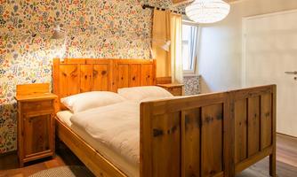 A cozy bedroom with a wooden bed and a colorful floral pattern on the wall. Next to the bed is a nightstand, and a warm light source creates a pleasant atmosphere.