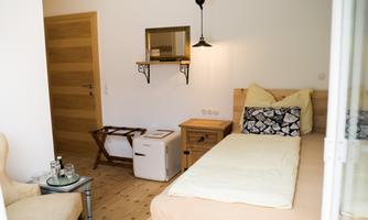 A cozy room with a single bed and modern wooden interiors. There is a small table, a chair, and a corner with a mirror.