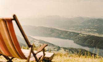 A cozy lounge chair is situated on a meadow with a view of a clear lake and the surrounding mountains. The landscape radiates tranquility and relaxation.