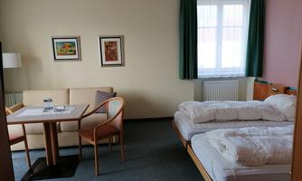 A simple hotel room with two single beds, a table, and two chairs. On the wall, there are some pictures, and there is a window with green curtains.