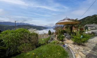 In front of our house is a green meadow, and a terrace with a light natural stone floor; in the background stands a stone fountain with a basin. From here, one looks out over the lake and the surrounding mountains.