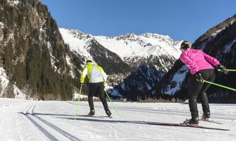 Two cross-country skiers in the snowy mountainous landscape. In the background, there are tall mountains and a clear blue sky.