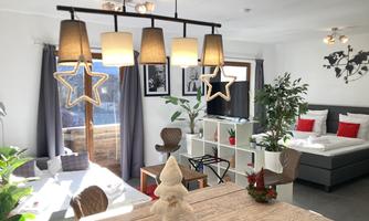 A cozy living room with a large dining table, modern furniture, and a clear view of a terrace. The decoration is simple, with plants and a festive Christmas tree.