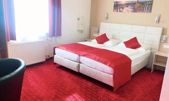 A modern hotel room with a large, comfortable bed and red accents. The room has a window with plenty of natural light.