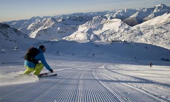 A skier glides over a perfectly groomed slope in the snow. In the background, impressive mountains can be seen under a clear sky.