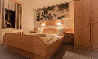 A cozy bedroom with a wooden bed and two pillows. A large-format picture of mountains hangs on the wall.