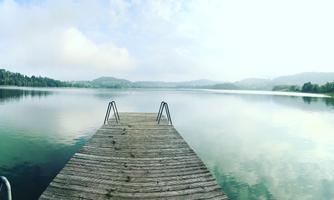 A wooden path leads to a peaceful lake. The landscape is surrounded by gentle hills and a cloudy sky.