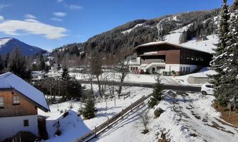 A winter landscape with snow-covered hills and trees. In the foreground, wooden houses and a modern building are visible.