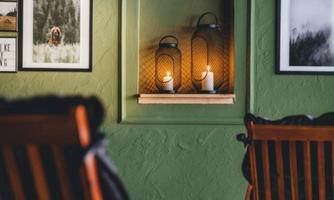 A cozy room with a green wall, two wooden chairs, and a decorative wall shelf. On the shelf are two lanterns and candles, while pictures hang on the wall.