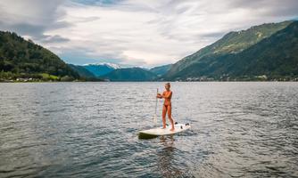 A person is standing on a paddleboard on a calm lake. In the background, green mountains and a cloudy sky can be seen.
