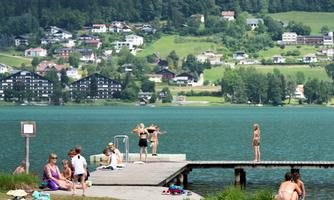 A tranquil lake with a wooden platform and several people relaxing and swimming. Surrounded by green hills and residential houses.