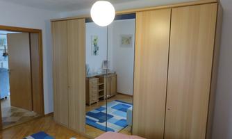 A bright bedroom with a large wardrobe, a table, and a colorful carpet. The room looks neat and inviting.