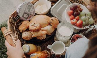 A basket full of fresh baked goods, fruit, and drinks. A person is holding the basket and enjoying a stylish picnic.