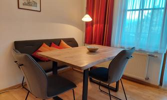 A cozy dining area with a wooden table and modern chairs. In the background, red curtains hang and a lamp provides warm light.
