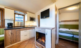 A modern kitchen with wooden details and a cozy fireplace. A bedroom is visible through an open room window.
