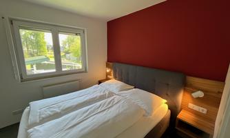 A modern bedroom with a gray bed and white bedcovers. A large window front offers a view of the outside.
