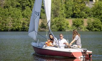A group of people is happily sailing on a boat on calm waters. In the background, green trees and a sunny landscape can be seen.