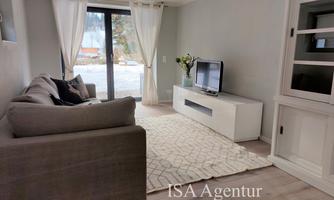 A bright living room with a gray sofa and a white TV unit. Large windows let in plenty of light and offer a beautiful view outside.