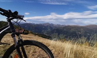 A mountain landscape with a view of the valley and the distant mountains. In the foreground, there is a mountain bike next to a backpack.