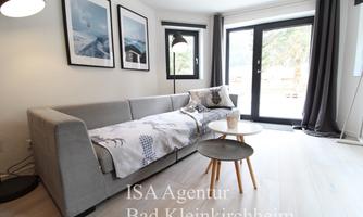 A modern living room with a gray sofa and a small table.  
Large windows provide plenty of daylight and offer a view outside.