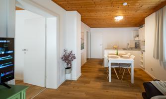 A modern and bright living and dining room with a wooden ceiling. The open kitchen is well-equipped, and a television is in the room.